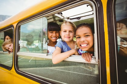 Elementary school students smiling out of a yellow school bus window